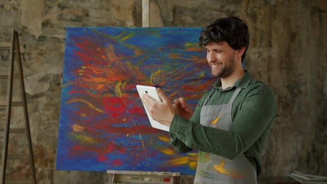 Male artist standing in his studio using a tablet wins. Workshop with oil painting