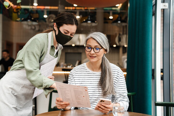 Fototapeta na wymiar Young waitress woman wearing face mask showing menu to her client