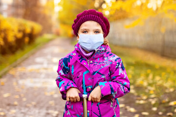 Girl of 3 years in purple clothes, protective mask with scooter in autumn on street