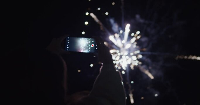A woman recording a video of exploding fireworks at night during a holiday celebration. 