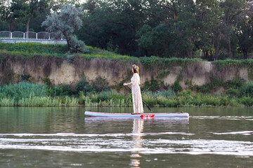 A beautiful woman in a dress with a wreath on his head, rowing on stand up paddle board (SUP) near shore at the river at summer morning on the holiday of Ivan Kupala