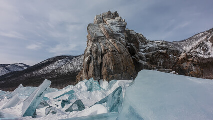 A bizarre rock, devoid of vegetation, against the backdrop of a blue sky and a wooded mountain range. Sheer slopes, flat top, cracks in rocks. In the foreground is a block of turquoise ice hummocks.