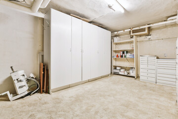 Various storage shelves and cabinet located on shabby floor near pipes and door in cellar