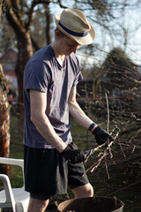 Young blond boy with a hat is chopping branches for later burning. Processing cut branches from fruit trees in orchards. Cutting branches into small sticks and putting them into a barrel.