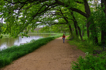 woman walks along a pedestrian alley in a park near a pond and among green trees. Natural background.