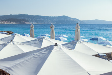 Obraz premium Sunny summer day and white umbrellas in Nice, France. Holiday atmosphere on summer time at French riviera. White umbrellas on a beach in blue clear sky and mountains on background. High quality photo