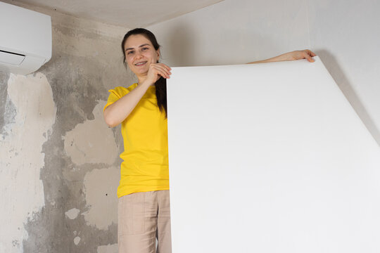 A woman repairman glues white wallpaper on the wall near the air conditioner under the ceiling.