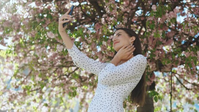 Young smiling woman in summer white dress taking selfie self portrait photos on smartphone. Model posing on park sakura trees background. Female showing positive face emotions.