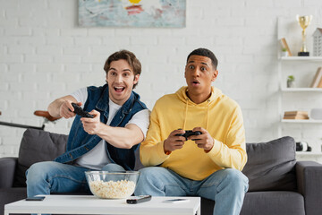 KYIV, UKRAINE - MARCH 22, 2021: interracial friends emotionally playing video game with joysticks