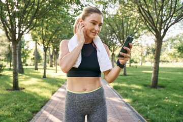 Use technology. Cheerful fit woman in sportswear and wireless earphones using her smartphone while jogging in a green park on a sunny day