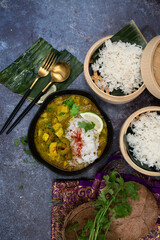 Vegan green Thai curry with squash and bitter gourd in bowl, steamed rice in bamboo steamers, banana leaf