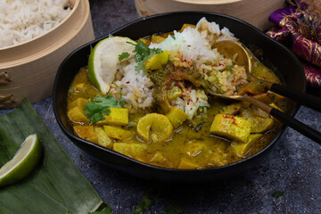 Vegan green Thai curry with squash and bitter gourd in bowl, steamed rice in bamboo steamers, banana leaf