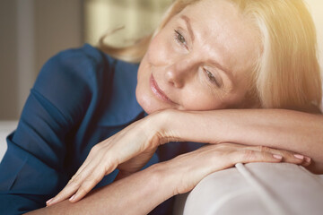 Close up portrait of attractive peaceful mature woman with pleasant smile relaxing on sofa at home