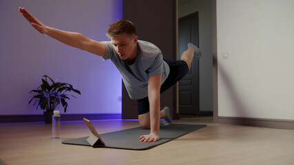 Skinny man doing morning exercises on yoga mat. He watches lessons on a tablet