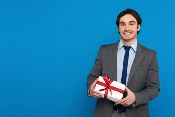 smiling young man in fashionable blazer holding gift box with red ribbon isolated on blue