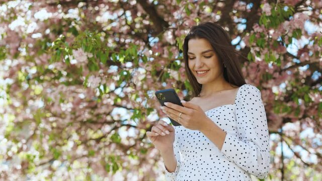 Smiling woman wearing white dress using smartphone. Girl using social media application text messages receive news smiling outdoor. Communication, social networks, online shopping concept. Technology