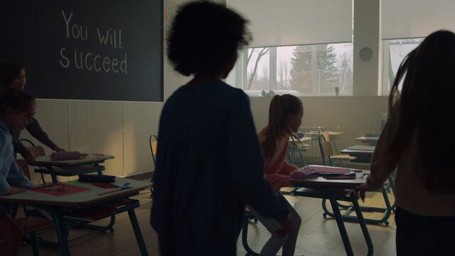 Students Walking In Classroom At School. Schoolchildren Sitting At Desks