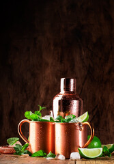 Moscow mule or mint julep alcoholic cocktail in copper mug with lime, ginger beer, vodka and mint. Wooden table, copper bar tools - obrazy, fototapety, plakaty