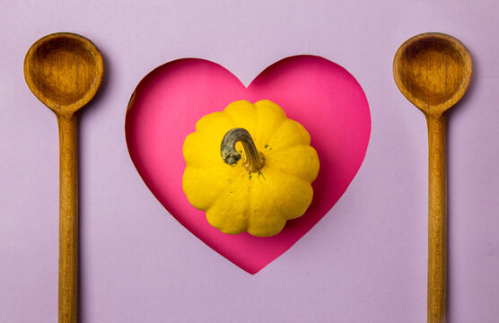 Pink heart shape on a purple background. In the heart is a small yellow pumpkin, and next to it are two wooden cooking spoons. Minimal concept of love and cooking.