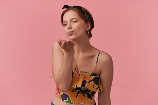 Image of european brown haired youth woman wearing yellow trendy summer clothes and black bandana emotion nice face wink looking at you lips kiss and send you air flying kiss against pink background