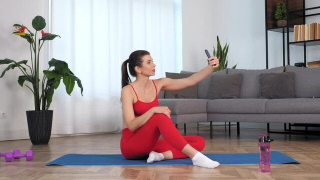 Smiling sportive woman in sportswear sitting on yoga blue mat greets records video on front camera smartphone. Positive fit sports girl shoots stories after fitness workout exercise training at home