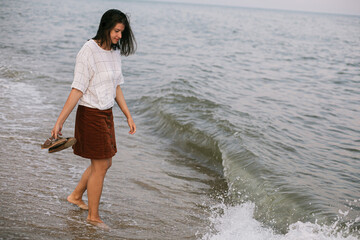 Carefree hipster woman walking barefoot on sandy beach and sea waves in the evening, enjoying calm...
