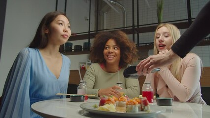 Cheerful attractive diverse multiethnic female friends deciding and choosing who will pay bill, playing rock paper scissors game during lunch at restaurant while waitress holding credit card reader.