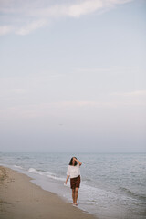 Carefree hipster woman walking barefoot on sandy beach with sea waves in the evening, enjoying tranquil moment. Casual young female  relaxing on seashore at resort. Mindfulness