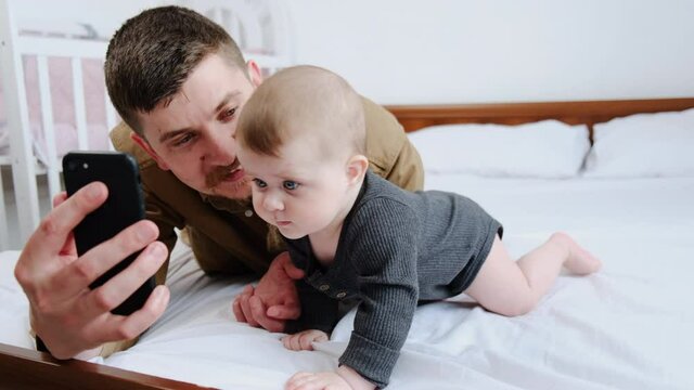 Close up of young father and cute beautiful newborn baby making selfie or video call to relatives lying on comfortable white bed at home. Technology, new generation, connection, parenthood concept