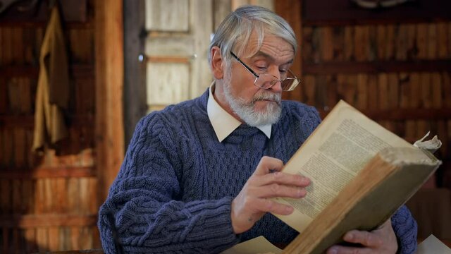 Old Gray Man Sitting At The Table And Reading Book. Image Of Wise Man With Beard. He Turns Pages.