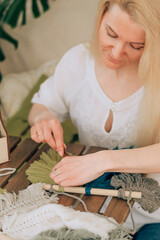 A young blonde woman weaving macrame in a home workshop.There are many indoor plants in the room.Handmade concept.
