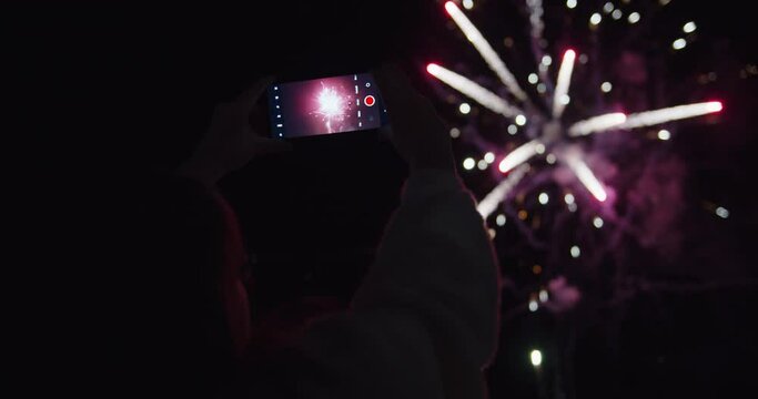 A woman in glasses taking a video of colorful fireworks at night sky.