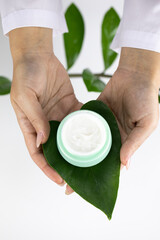 Close-up hands hold cosmetic jar with cream on fresh green leaf on white herbal background. Copy space. Plan-based cosmetics, skin care laboratory development, active natural ingredients concept