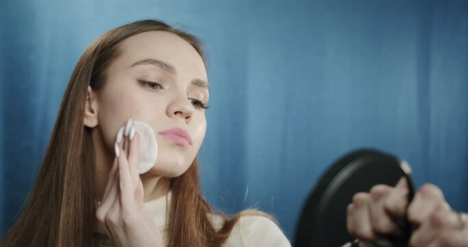Beautiful teenager washing off her makeup, wiping her face from cosmetic, using cotton pad, looking in mirror. Young woman posing in fashion studio on blue background. 