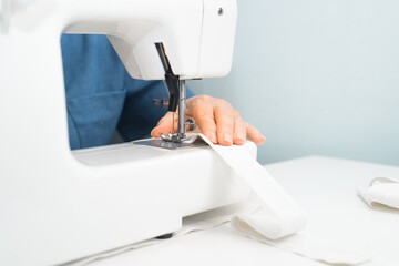 Woman working on sewing machine at home. Hands of senior dressmaker sews something from white fabric, close-up. Hobby or work at home for pensioner. Selective focus on hands