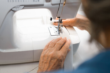 An elderly seamstress using sewing machine at home. Older woman is sewing something from a white cloth, view from the back. Hobby or work at home for a pensioner. Selective focus on hands