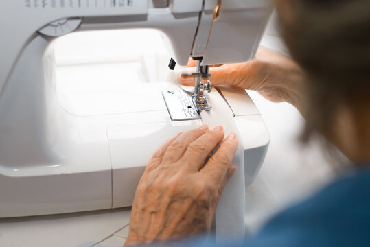 An Elderly Seamstress Using Sewing Machine At Home. Older Woman Is Sewing Something From A White Cloth, View From The Back. Hobby Or Work At Home For A Pensioner. Selective Focus On Hands