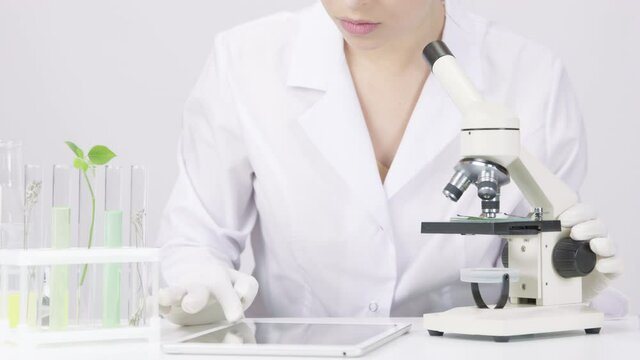 Close-up young attractive female microbiologist works on research, looks through microscope and records clinically proven results on tablet on white laboratory background. Science-based, healthcare