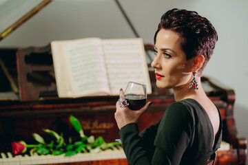 Side view portrait of attractive and elegant woman sitting at the piano and holding wine glass. Red rose on piano keyboard