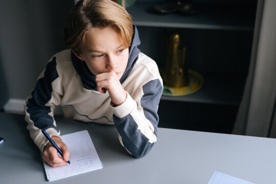 Close-up hands of unrecognizable pupil boy writing in paper notebook solving math equations sitting at desk in dark room. Back view of schoolboy doing homework at home.