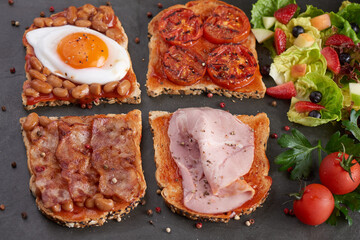 Variety of open sandwiches made of brown whole wheat  bread with tomatoes sauce, tomatoes, white beans, bacon, fried egg and smoked ham. Fresh fruit and vegetable salad