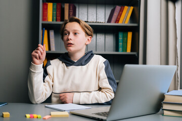 Portrait of surprised pupil boy having idea and raising hand during learning online using laptop at desk, looking away. Child schoolboy doing homework at home. Concept of remote online education.