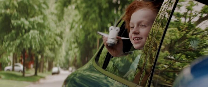 CU Portrait of cute little dreamer kid boy sticking head out of car window and flying his toy spaceship while riding through neighborhood. Shot with 2x anamorphic lens