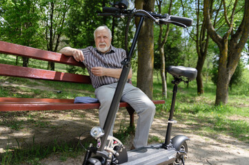 Fototapeta na wymiar A positive, elderly man went for a walk to the park, stopped and sat down to rest on a bench next to an electric scooter. Summer, sunny day. Healthy lifestyle of the elderly.