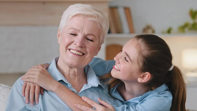 Happy relaxed granddaughter embracing older adult granny cuddling bonding rest on couch at home. Old grandmother touches nose child. Affectionate 2 two age generations family enjoy time together