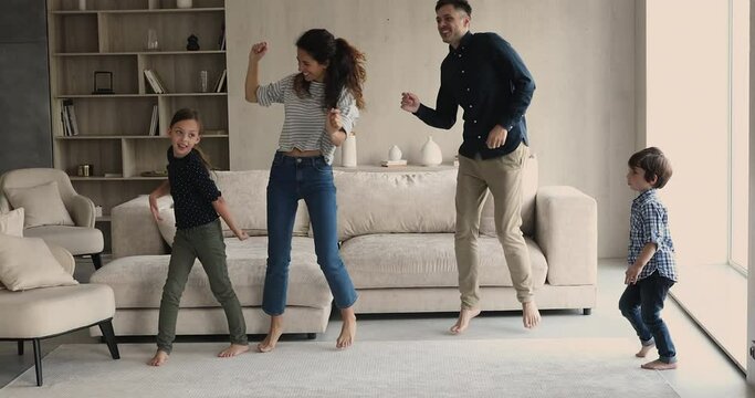 Happy homeowners family dancing barefoot in fashionable living room. Married cheery couple their beautiful active children listen to music funny moving at home, having fun on weekend. Hobby concept