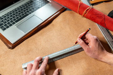 Point of view unrecognizable male taking measures with pencil and a ruler on a piece of leather with computer laptop.