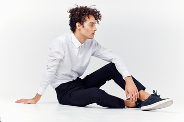 curly-haired guy in shirt and trousers and sneakers sits on the floor indoors side view