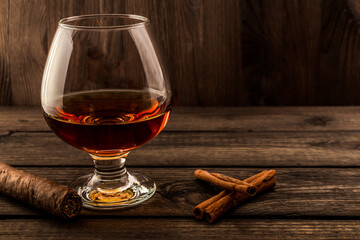 Glass of brandy with cinnamon sticks and cuban cigar on a old wooden table. Focus on the cinnamon sticks