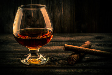 Glass of brandy and two different cuban cigars on a old wooden table. Focus on the cuban cigars